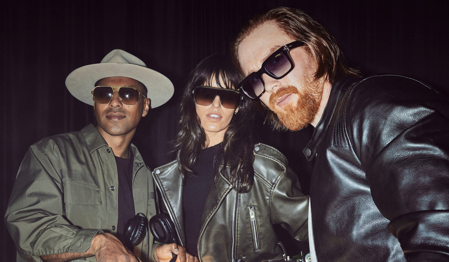 Three people wearing sunglasses and leather jackets pose confidently indoors.