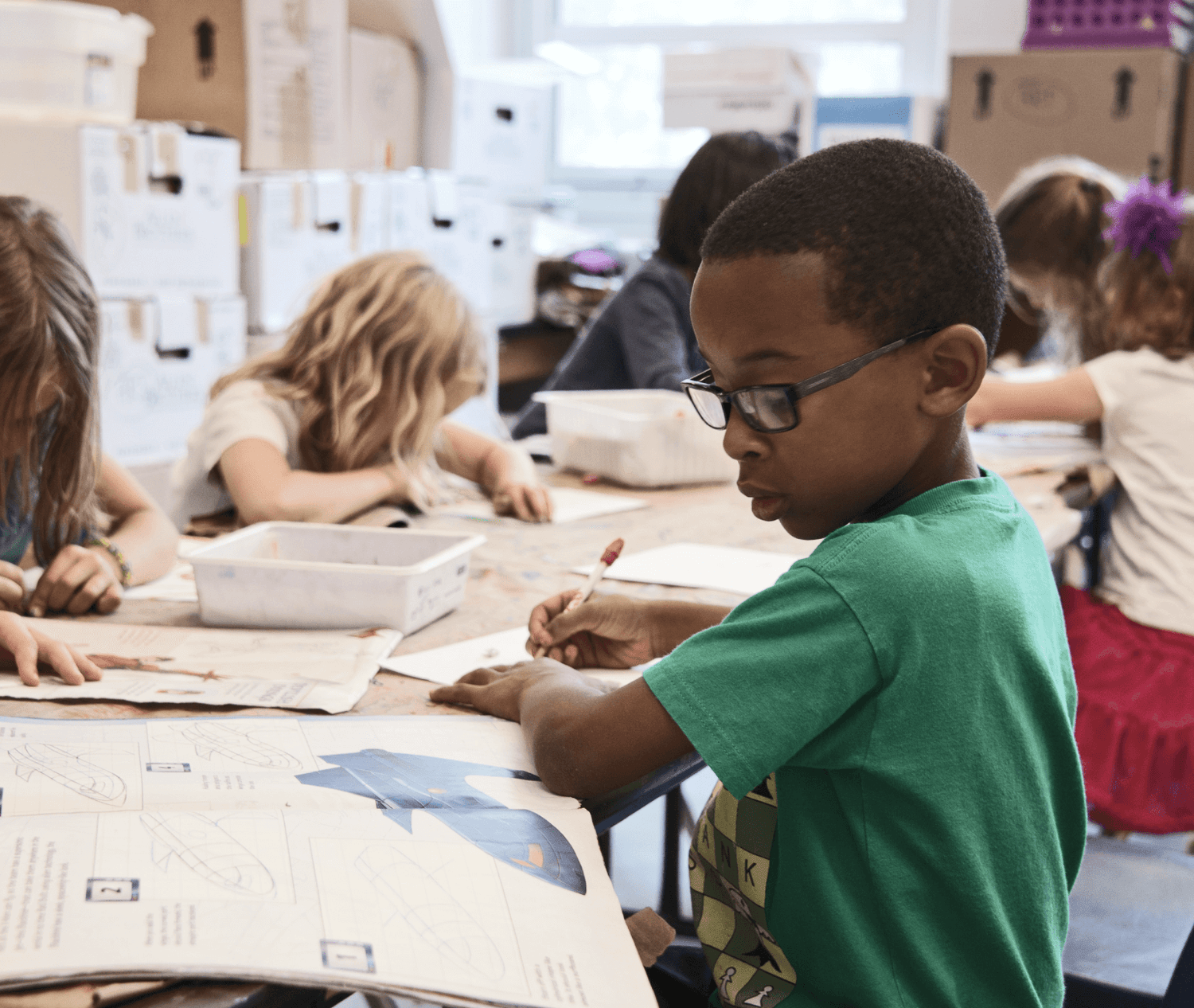 Several kids are sitting down and writing in a classroom. The lead kid is putting on glasses for better vision.