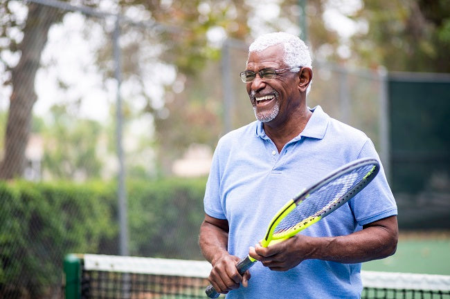 A senior is putting on progressive lenses and standing on the tennis court with a racket in his hand.